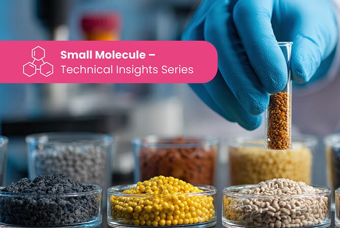 Blue-gloved hand holding a small glass vial of tan granules above an array of petri dishes filled with multi coloured powders and pellets in a laboratory setting.